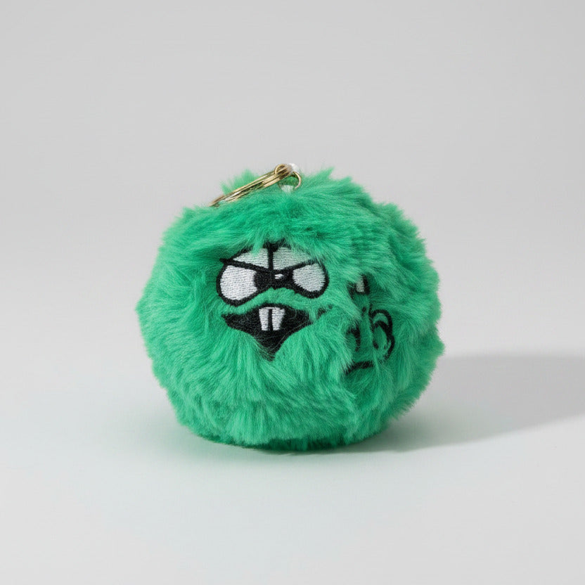 A green fuzzy plush keychain or bag charm with a cartoon face and black markings, lying on a brown wood-grain surface.
