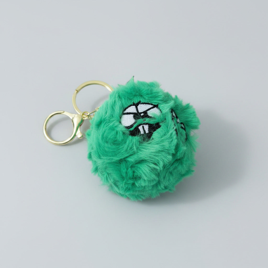 A green fuzzy monster keychain with a gold-tone split ring and clasp, featuring embroidered eyes and a mouth, resting on a wooden surface.